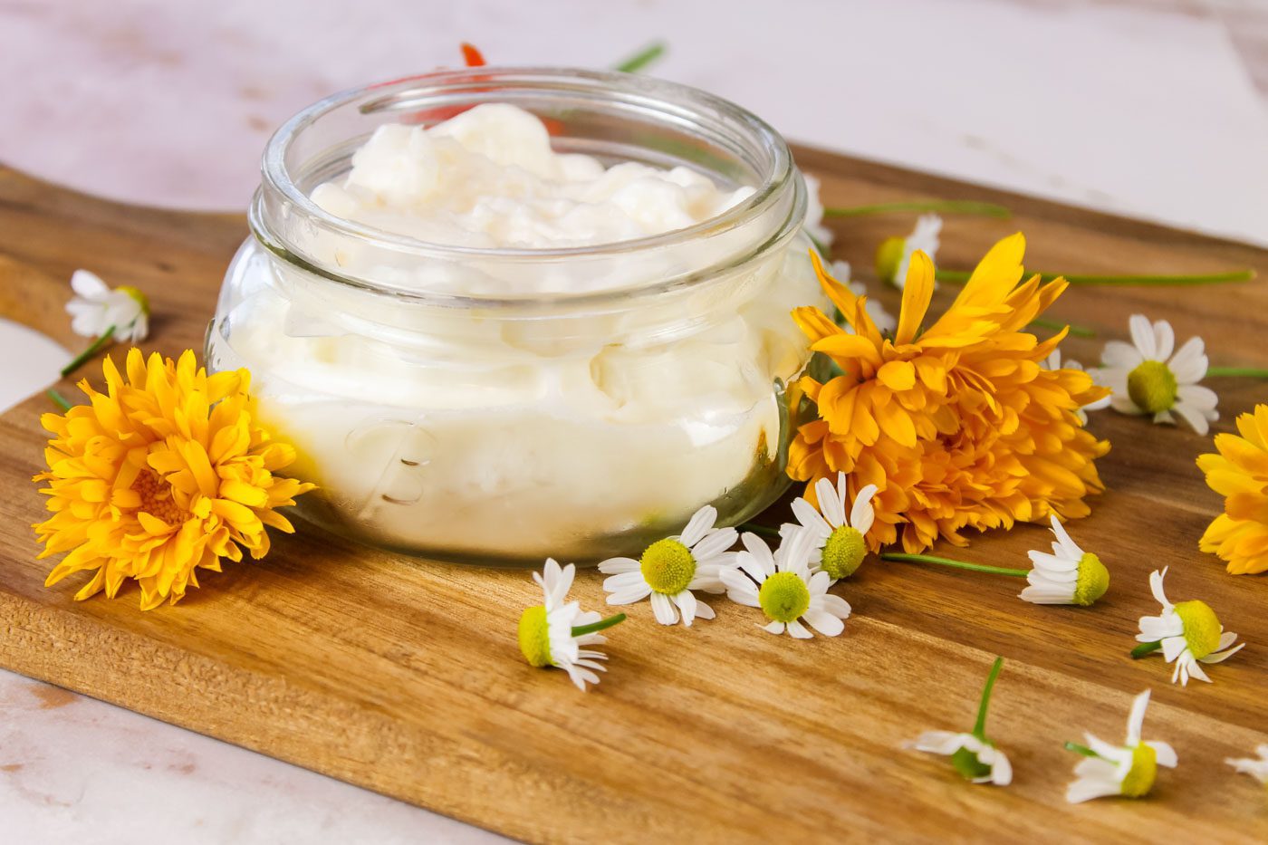 glass jar of lotion sitting on a wooden cutting board surrounded by calendula and chamomile flowers