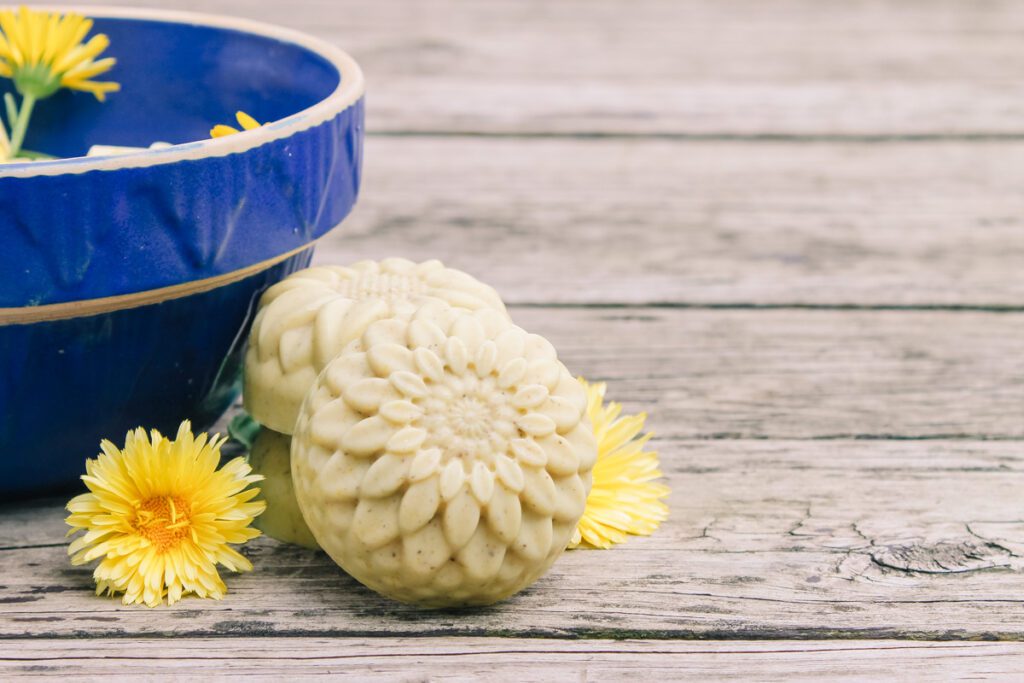 two bars of homemade soap sit next to a blue bowl and some fresh flowers