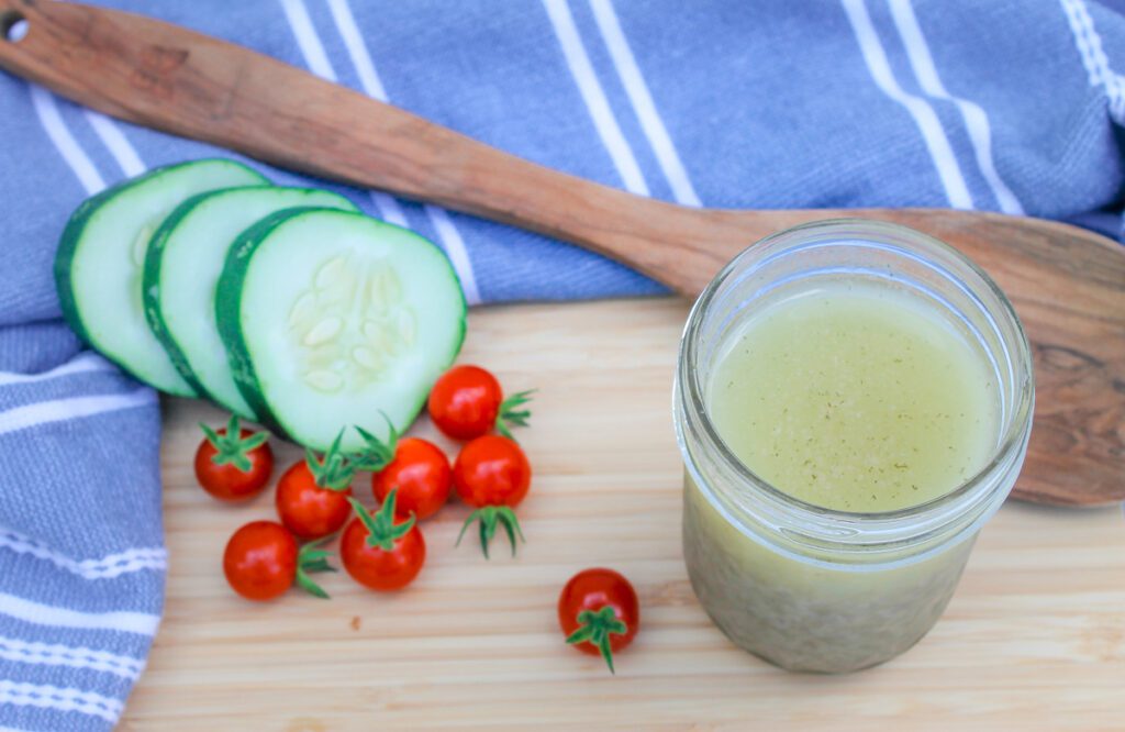 top down view of a mason jar filled with salad dressing next to a wooden spoon and some sliced cucumbers and tomatoes