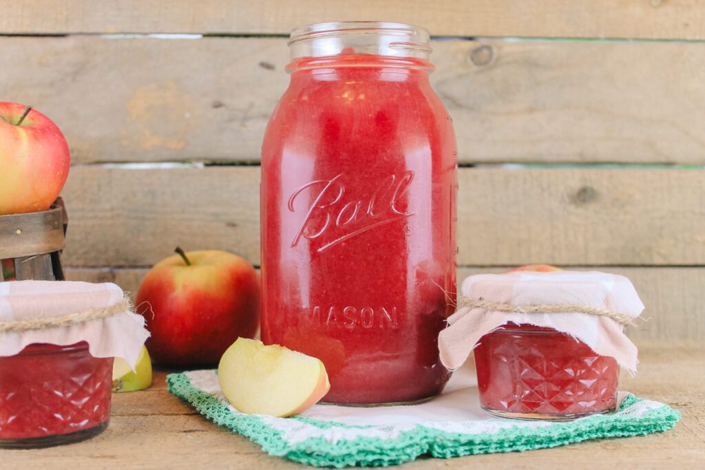 large jar of homemade strawberry applesauce sitting beside a couple small jars of sauce and some sliced apples