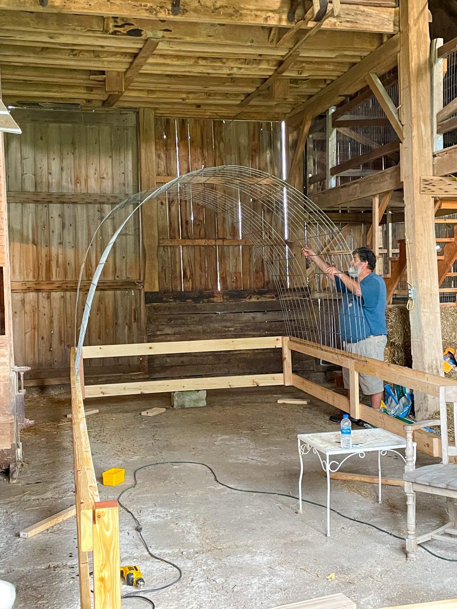 inside a barn a man bends a cattle panel to create the frame for a diy greenhouse