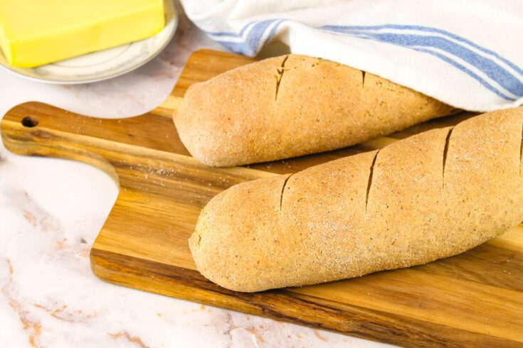 two loaves of easy french bread with fresh milled flour sit on a wooden cutting board beside a stick of butter