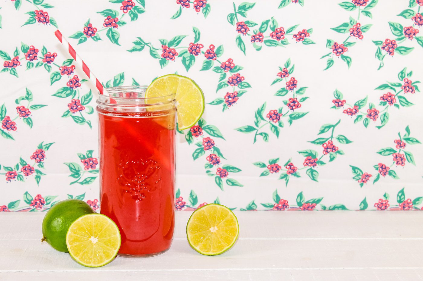sitting in front of a floral tablecloth is a mason jar full of cherry limeade probiotic soda