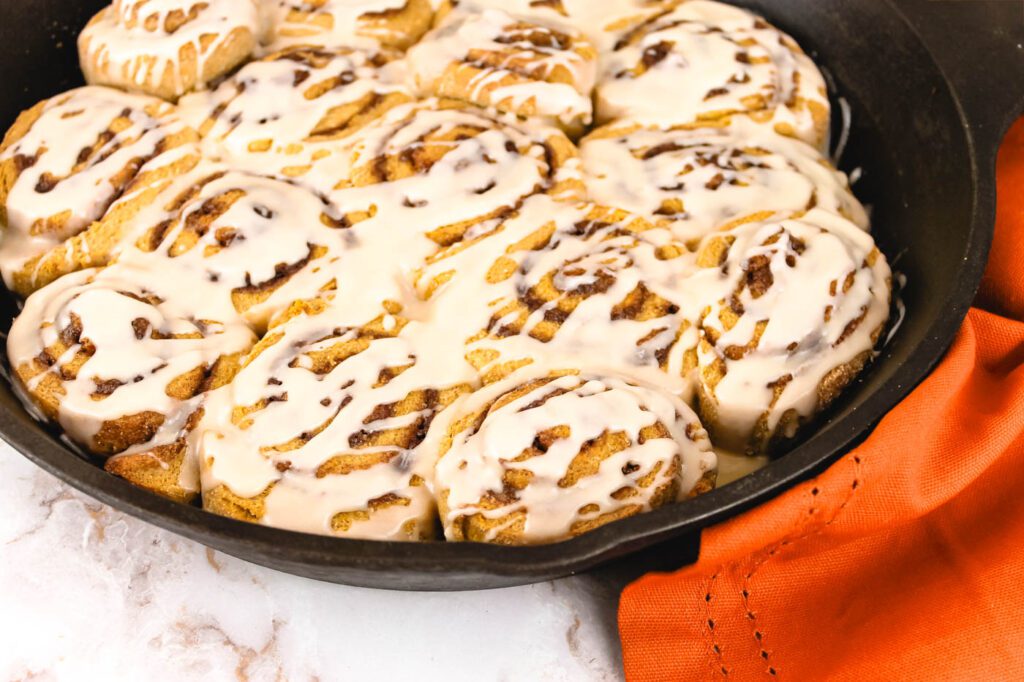 an orange tea towel surrounds a cast iron pan filled with cinnamon buns