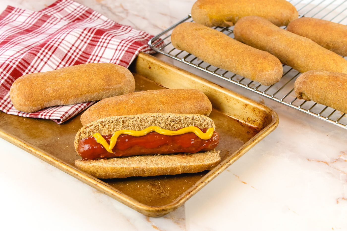 several hot dog buns sit scattered on a kitchen countertop, one is slit open and filled with a grilled hot dog with ketchup and mustard