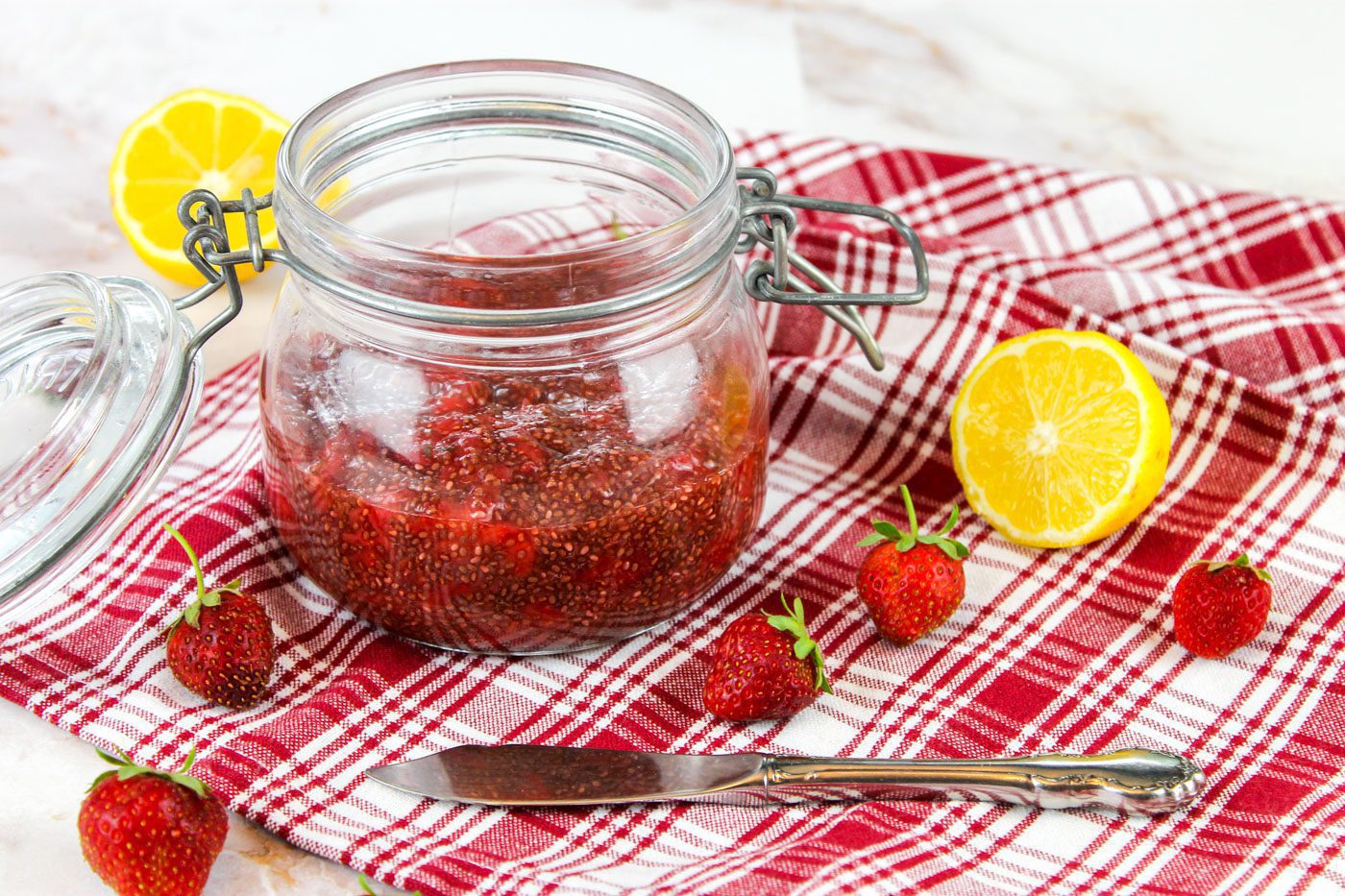 glass jar full of jam sits on top of a red plaid towel covered in strawberries and lemons