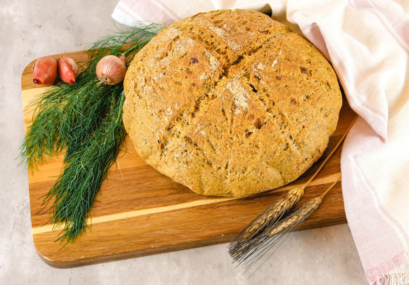 large loaf of bread sits on a cutting board surrounded by herbs and onions