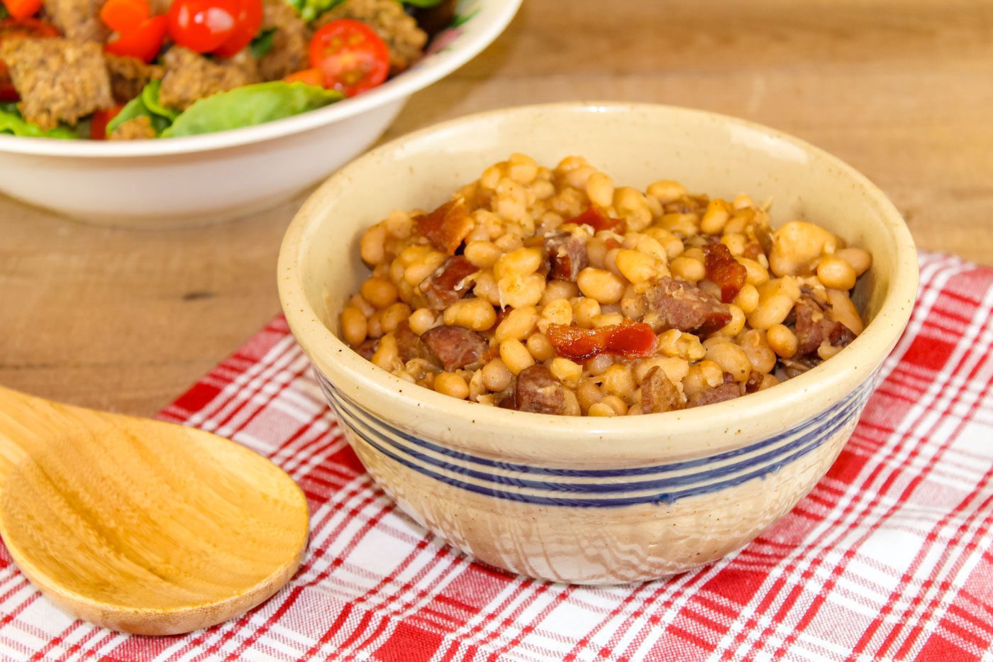 small bowl of homemade baked beans sits on top of a plaid napkin next to a wooden spoon