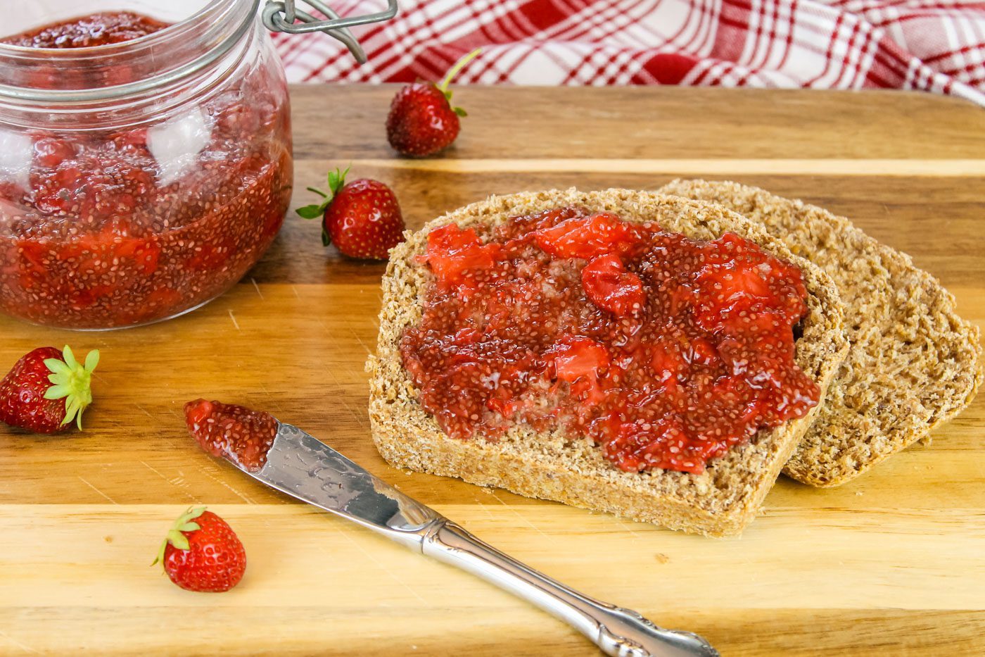 a slice of bread covered in jam sit next to fresh strawberries and a jar of chia seed jam