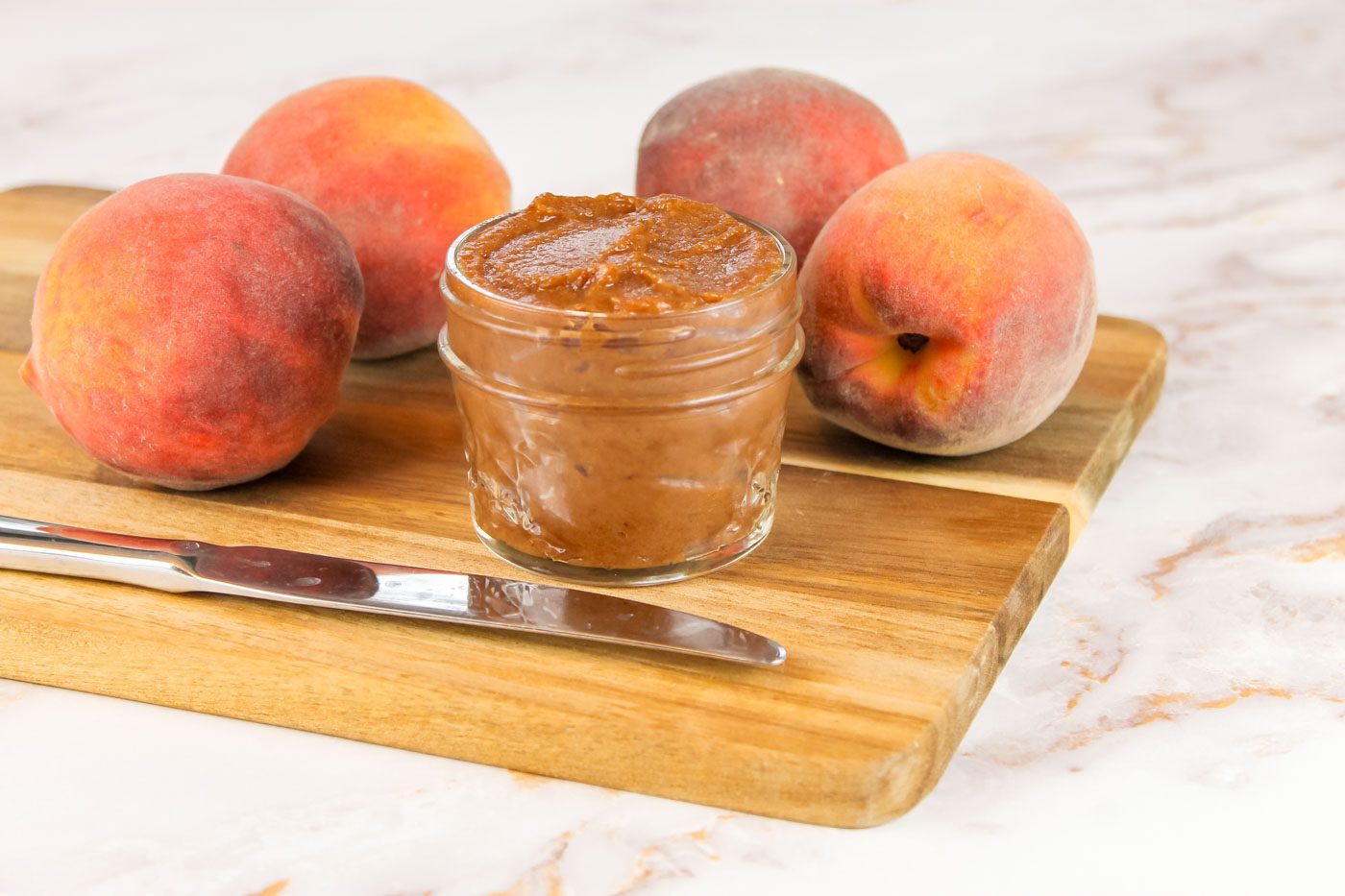 on a cutting board sits a butter knife, peaches and a glass jar of peach butter