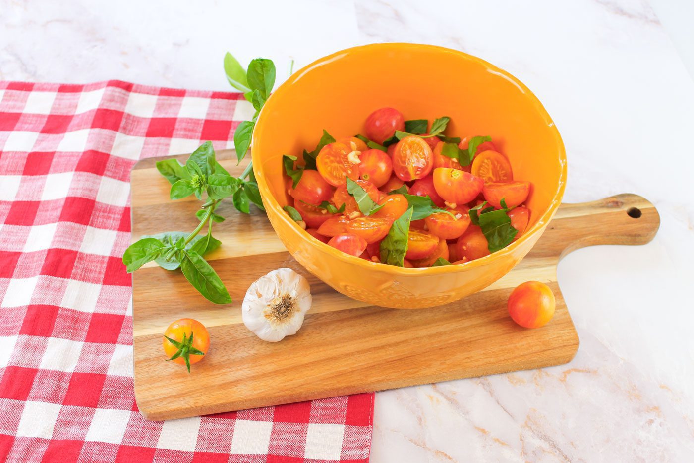 on top of a red plaid napkin is a cutting board and yellow bowl of sliced tomatoes, garlic and shredded basil