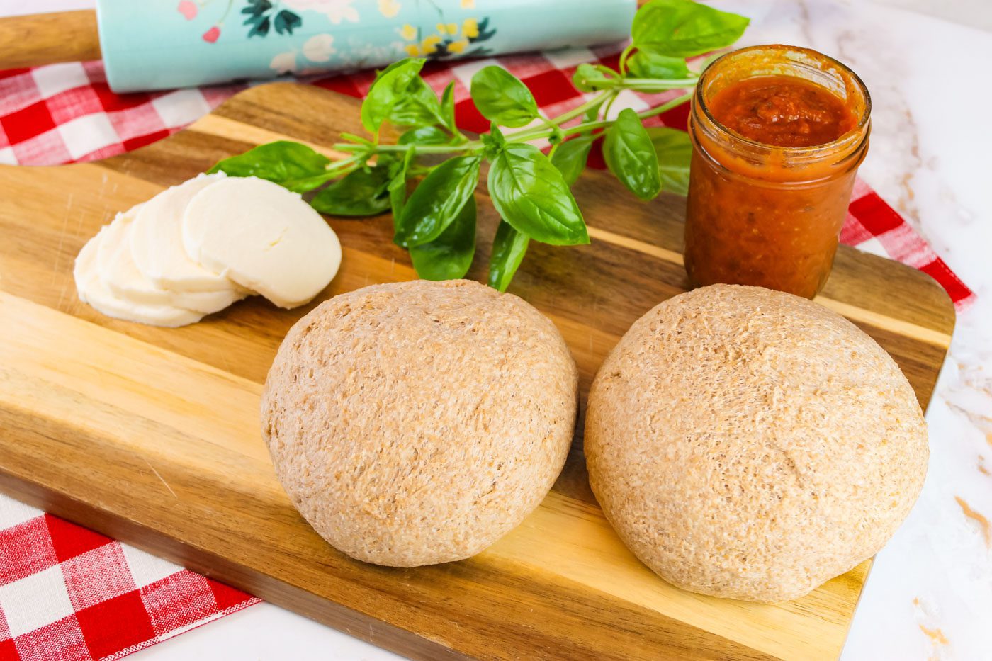 two balls of dough sitting on a wooden cutting board. Board has slices of mozzarella cheese, fresh basil and a jar of pizza sauce.