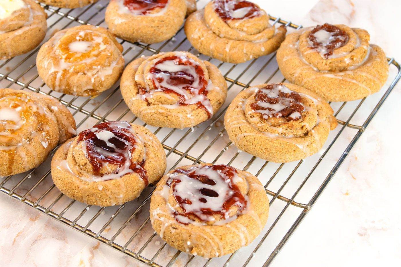 a dozen danishes filled with different jams sit on a metal cooling rack against a marble countertop