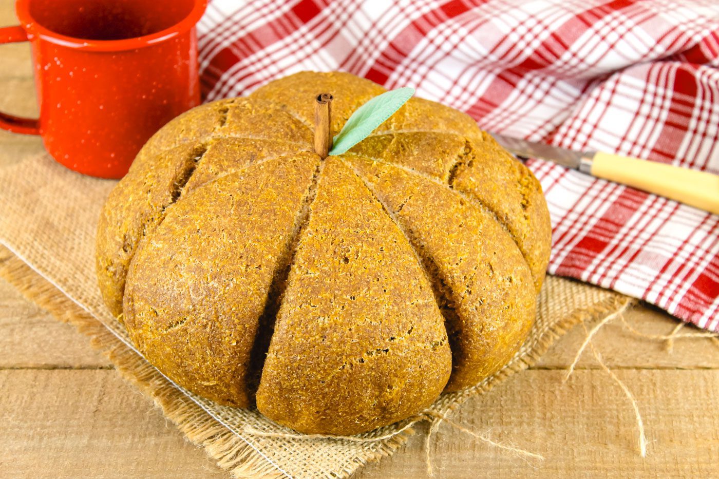 pumpkin shaped loaf of bread sits in front of a red mug and a red plaid kitchen towel