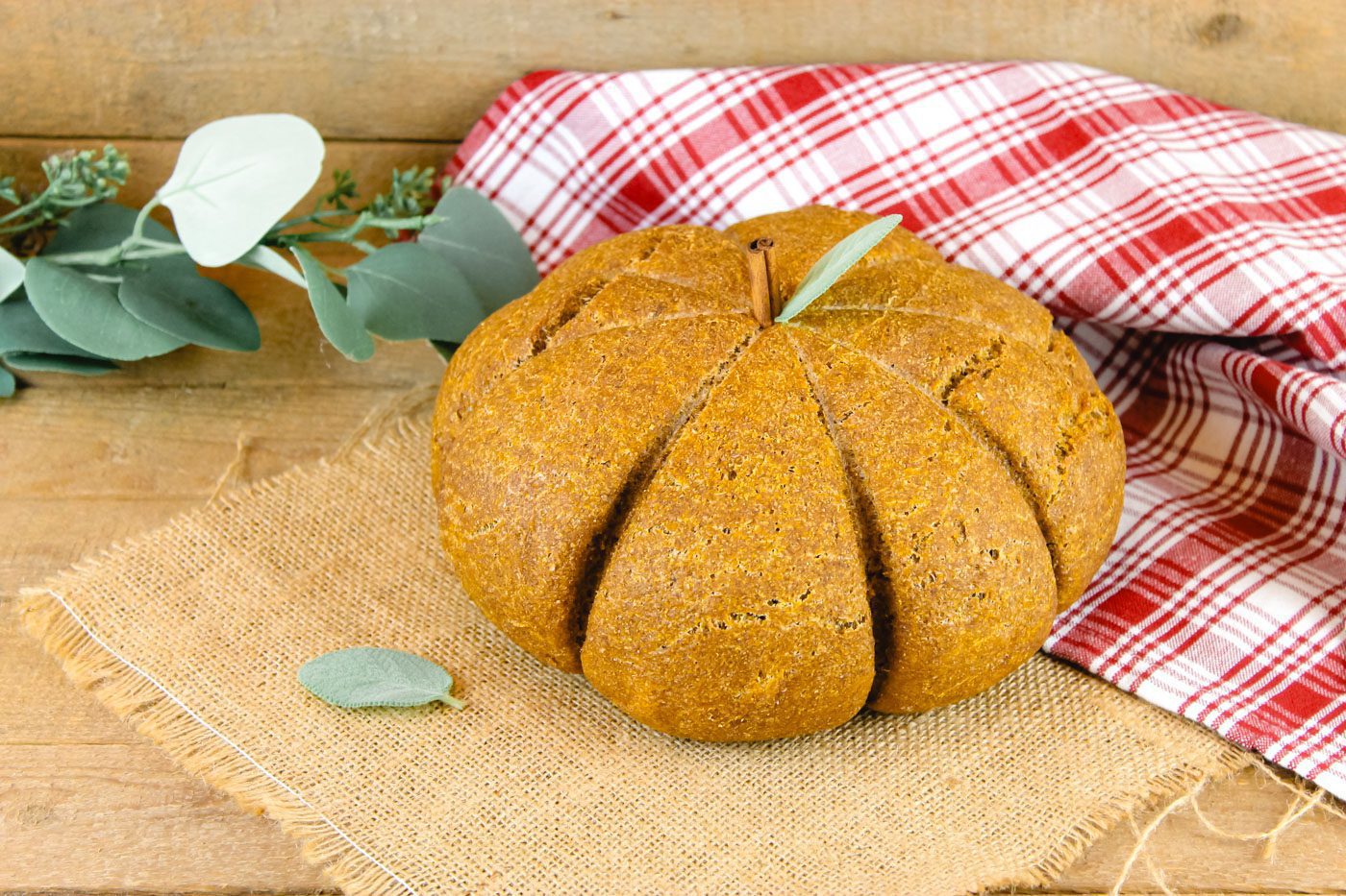 top view of a pumpkin loaf set against a red plaid background, green sage leaves and some burlap set as a tablecloth