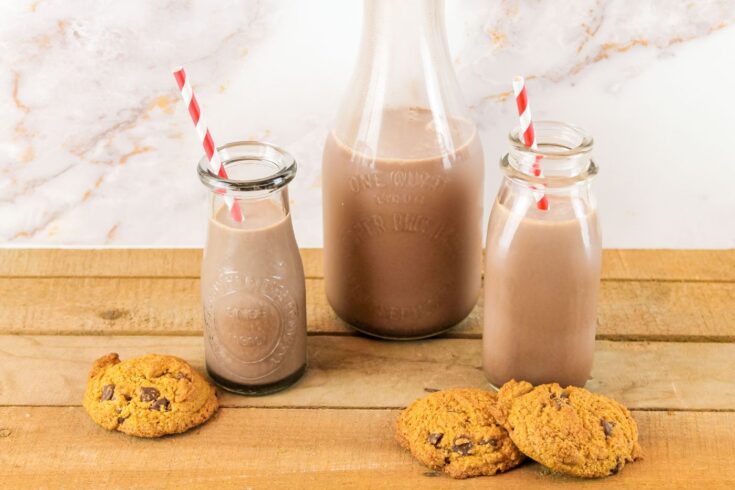 3 chocolate chip cookies sit in front of 3 bottles of chocolate milk. two bottles have red and white straws coming out of the top of the bottle.