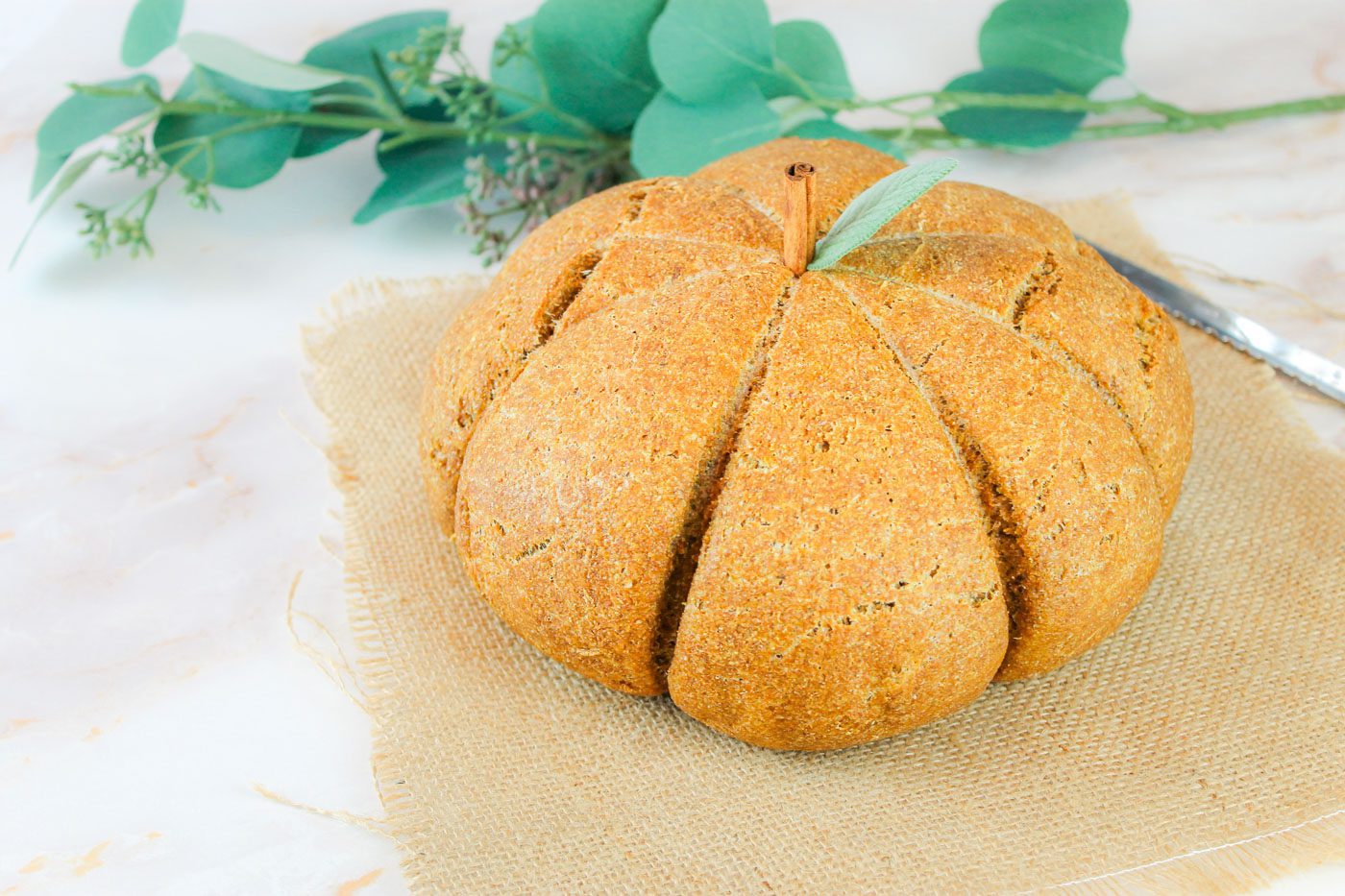 a loaf of bread shaped like a pumpkin sitting on a piece of burlap and a branch of greenery in the background