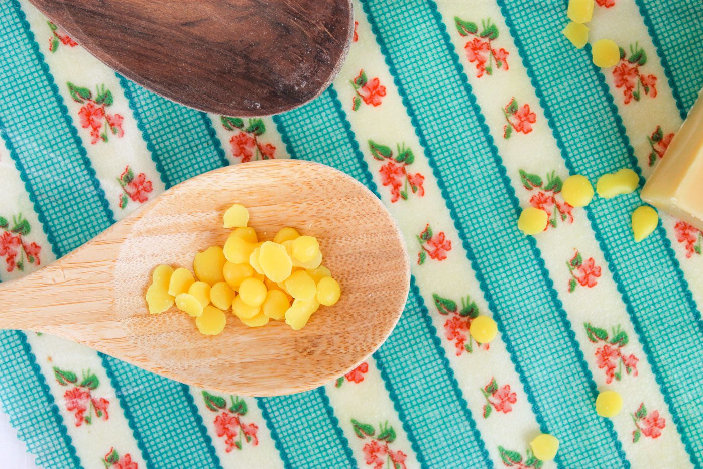 a wooden spoon full of beeswax pellets sits on top of a floral piece of fabric