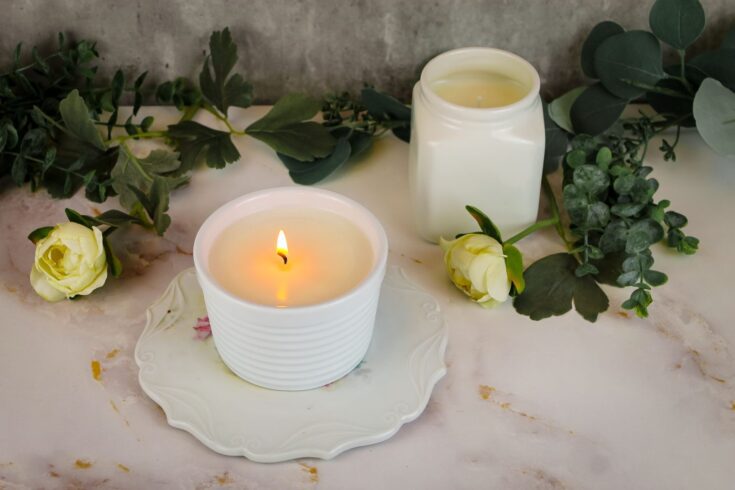 two candles, one lit sitting on a marble countertop surrounded by white roses and greenery