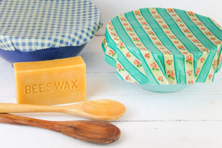 on a white wooden background sets two bowls covered in beeswax wraps, a block of beeswax and two wooden spoons