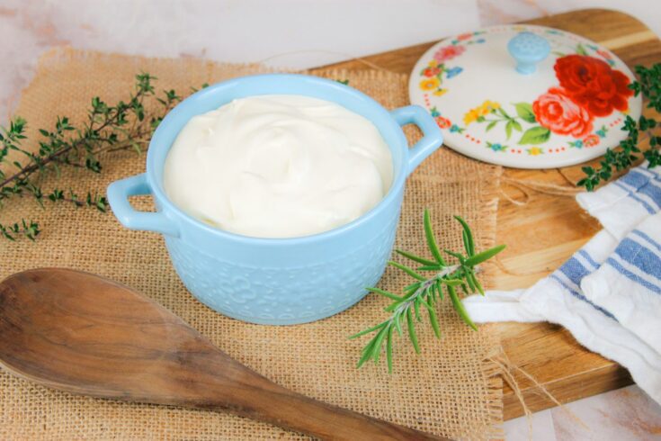 bowl of sour cream sits on a wooden cutting board surrounded by green herbs