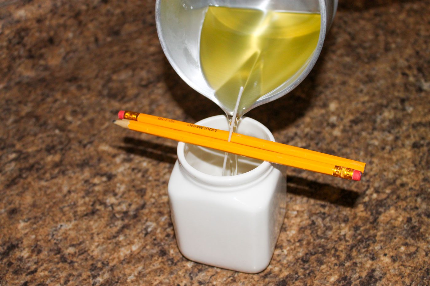 melted candle wax pouring in a white container. two pencils set across container to hold candle wick in place.