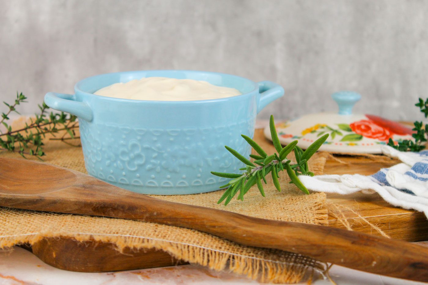 blue container full of sour cream sits on a wooden cutting board that has herbs and a towel and a wooden spoon