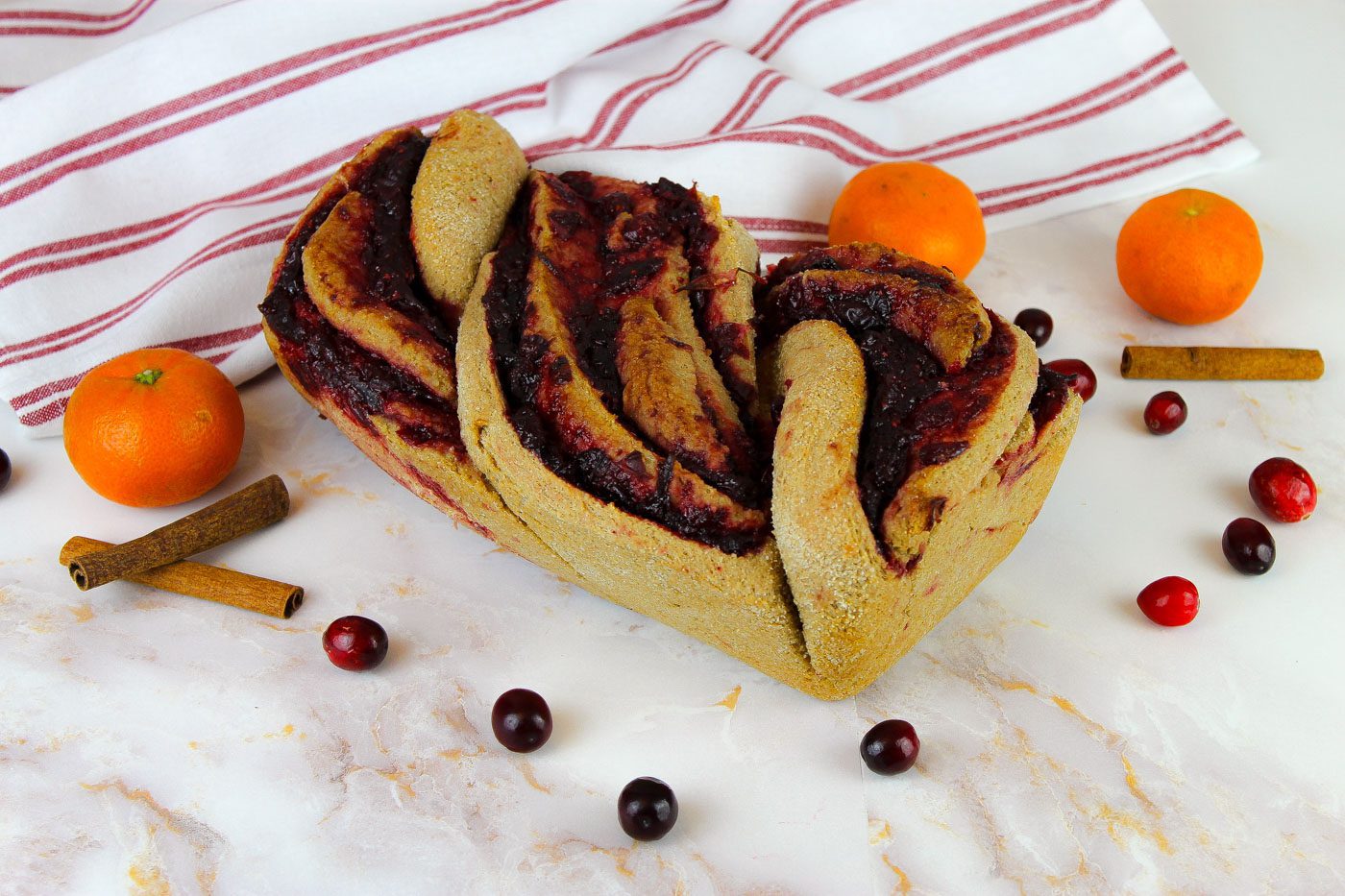 loaf of cinnamon bread surrounded by a red striped tea towel, oranges and cranberries