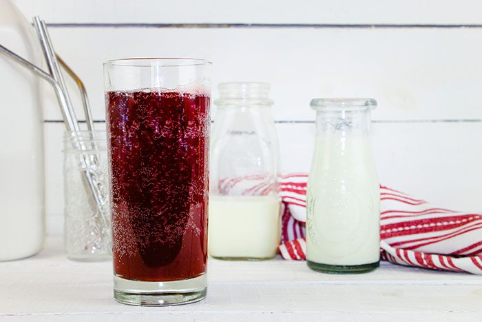 glass of cherry kefir soda sits besides a couple of milk bottles and a red striped towel