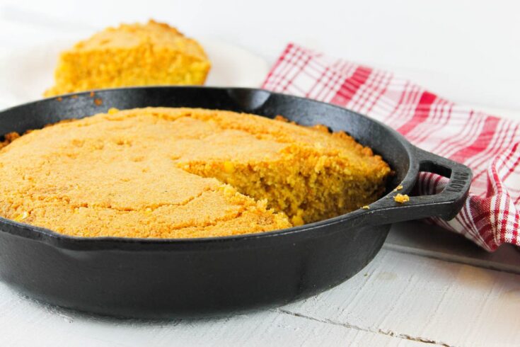cast iron skillet filled with cornbread sits on a wooden table surrounded by a tea towel and a slice of cornbread