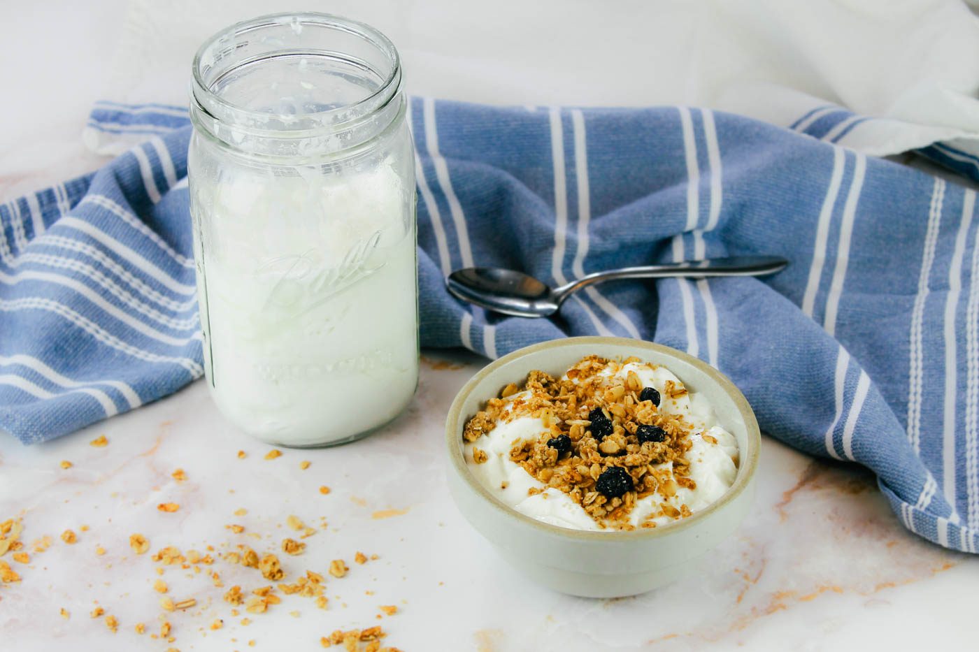 glass jar of yogurt sits beside a blue kitchen towel and a bowl of yogurt and granola