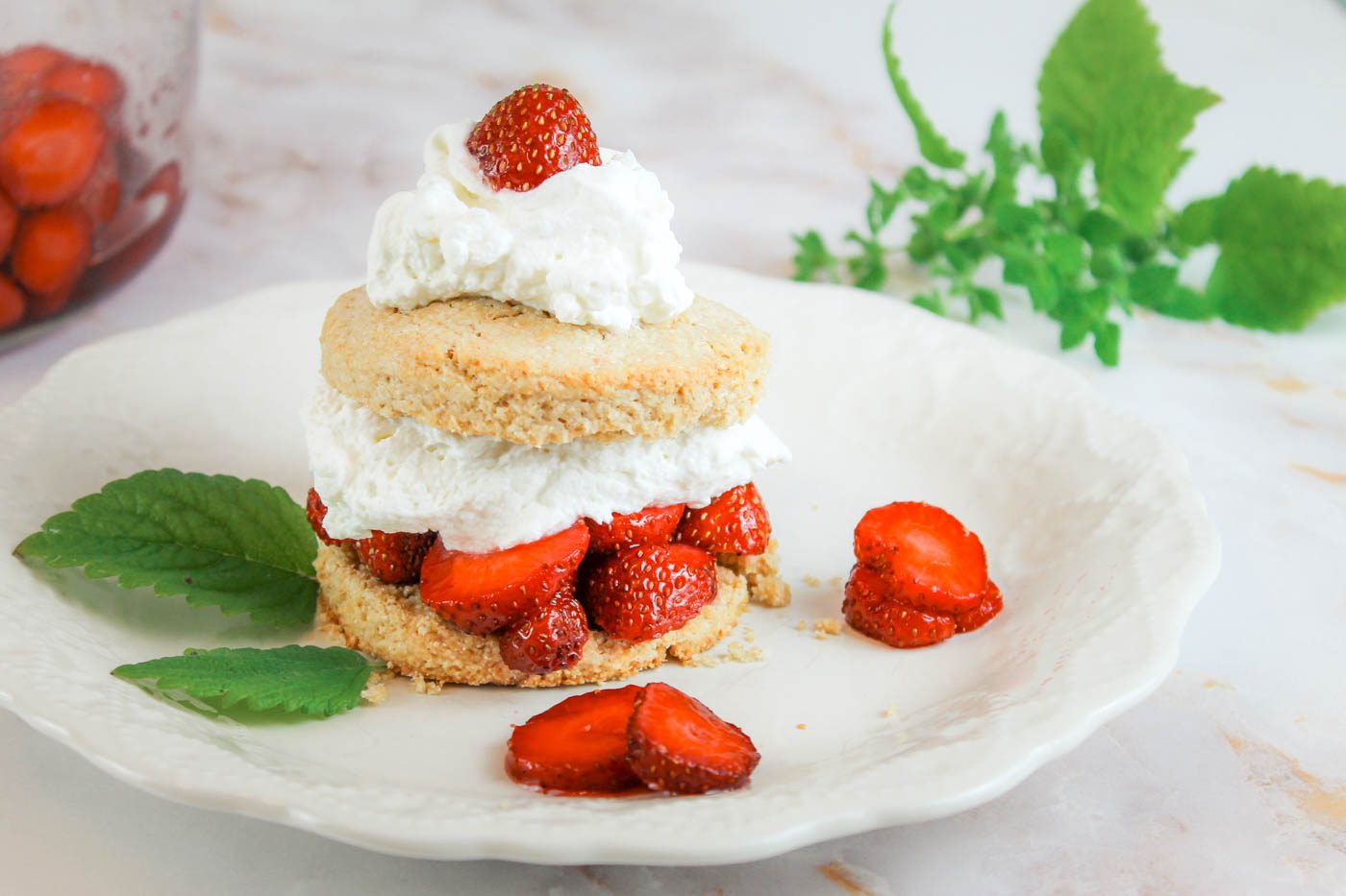 a white plate with a layered strawberry shortcake. in the background is a bowl of sliced berries