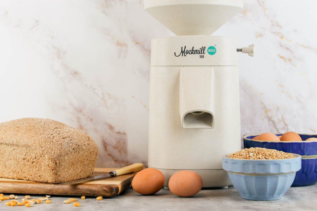stone mill sitting next to a bowl of eggs, a loaf of bread and a small blue bowl of wheat berries