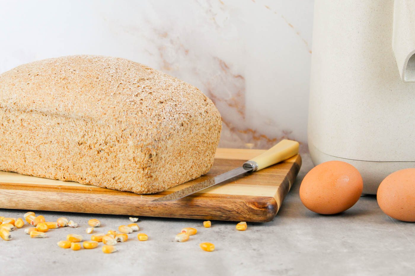 loaf of homemade bread sits on a wooden cutting board next to some dried corn, a knife and farm fresh eggs