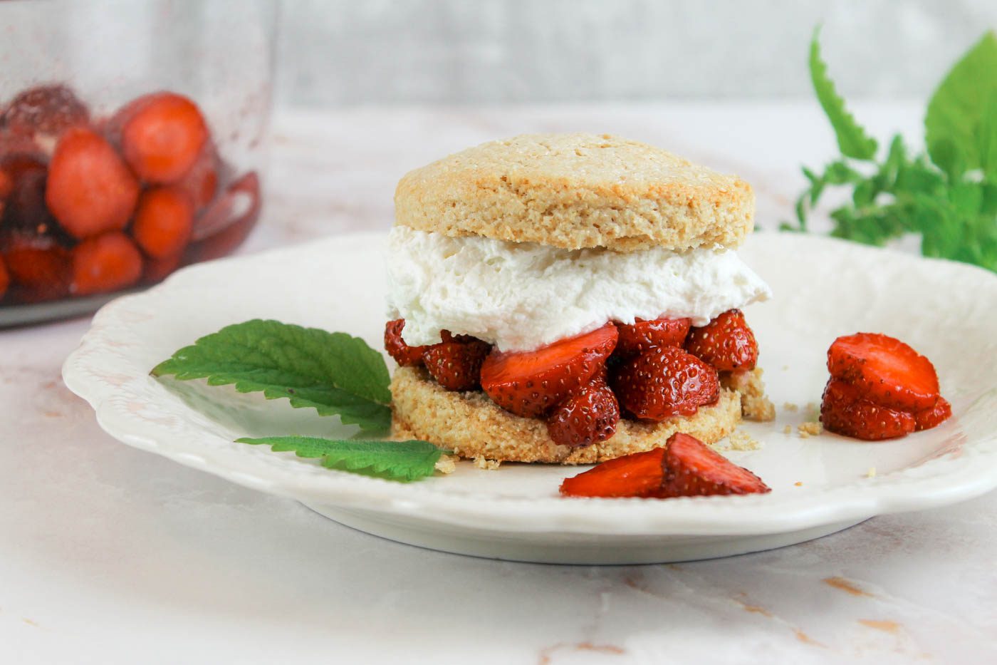 fresh milled flour biscuit filled with fruit and whipped cream sit on a marble countertop next to some herbs and a glass bowl of sliced fruit