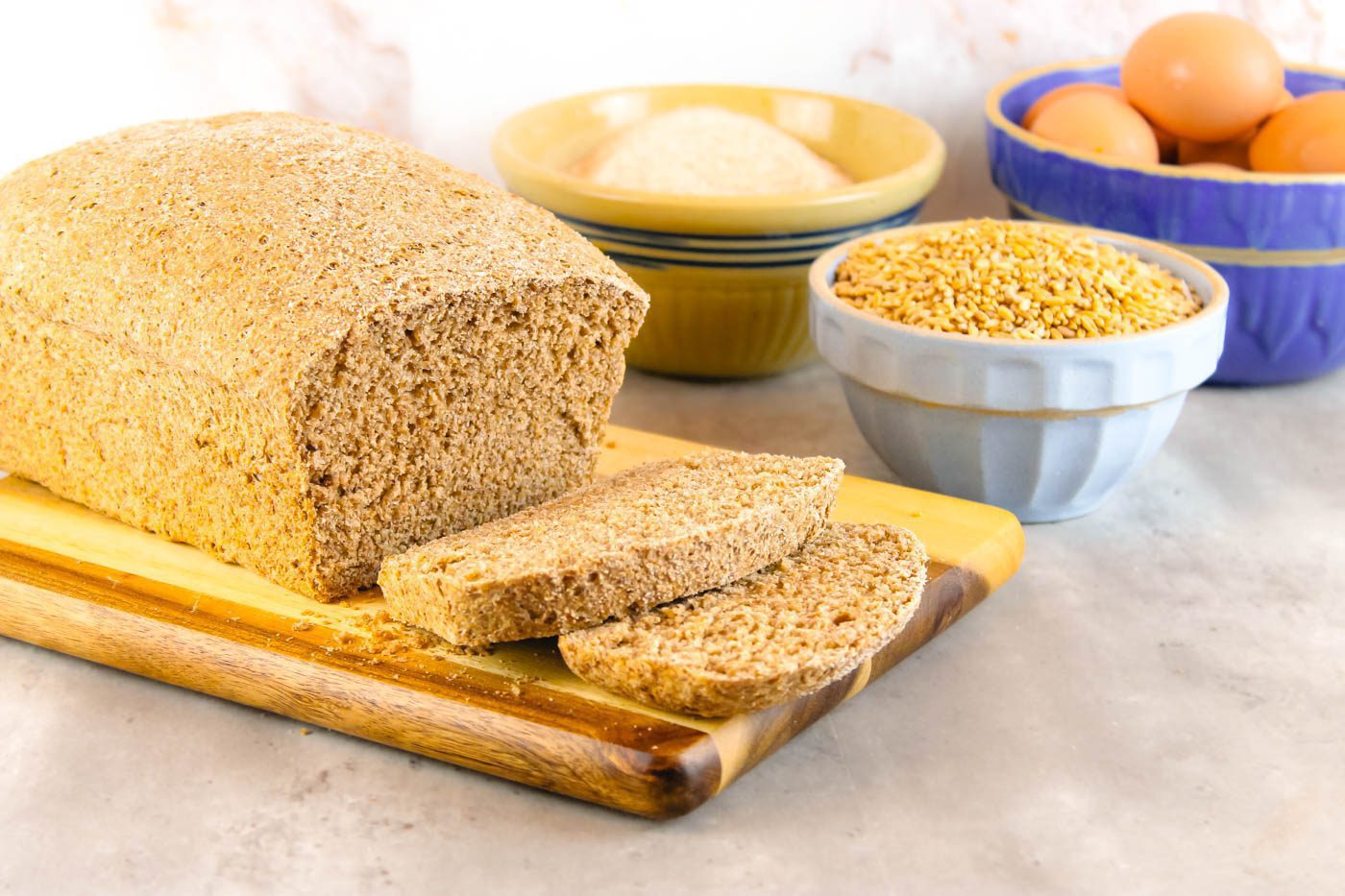 loaf of sandwich bread sits on a cutting board beside bowls of baking ingredients