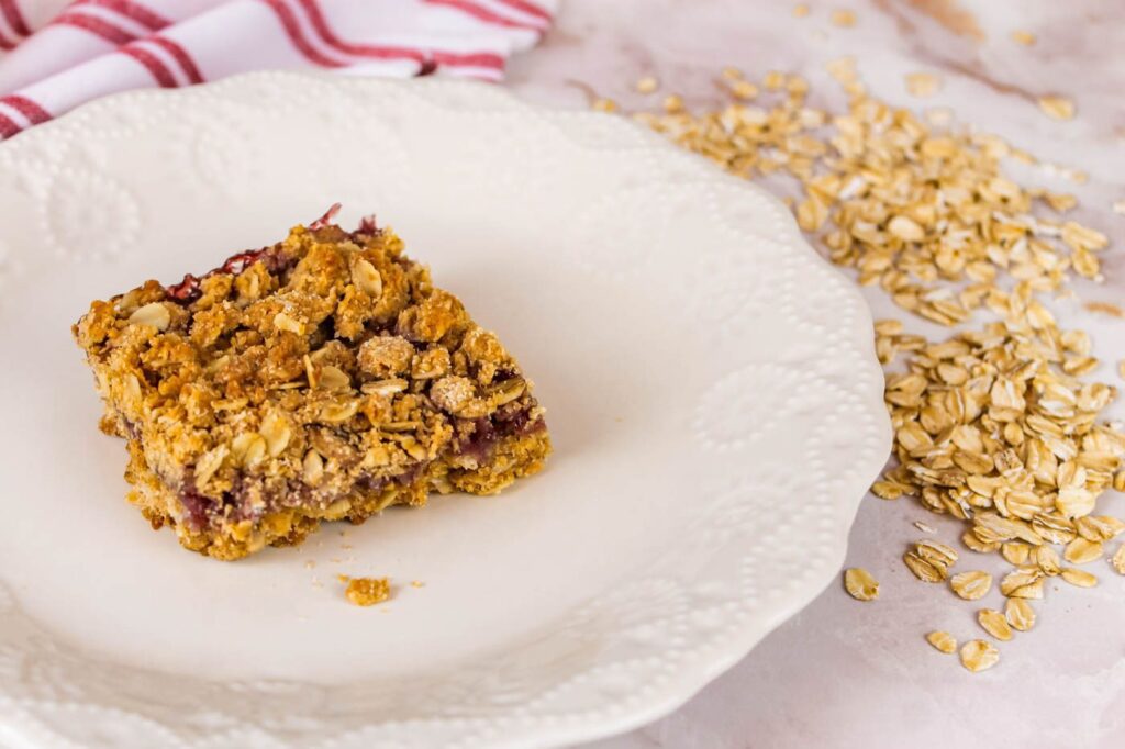 whole grain oatmeal jam bar sitting on a white plate next to a pile of oats and a red striped tea towel