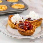 3 mini dutch baby pancakes filled with cherries and whipped cream sit on a plate in front of a muffin tin