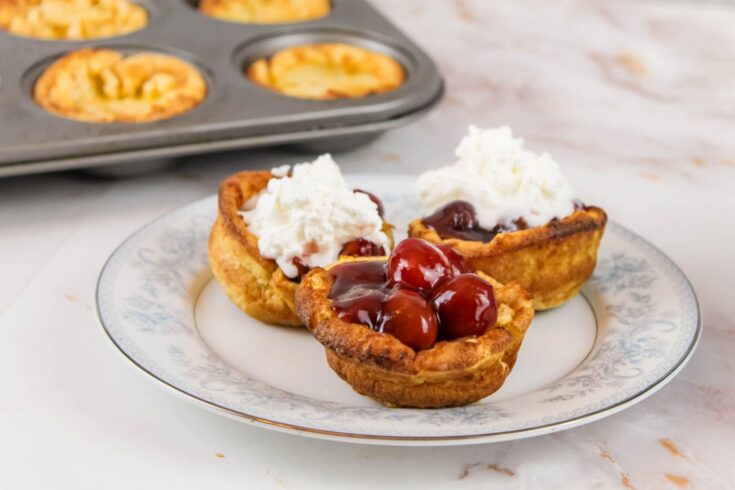 3 mini dutch baby pancakes filled with cherries and whipped cream sit on a plate in front of a muffin tin