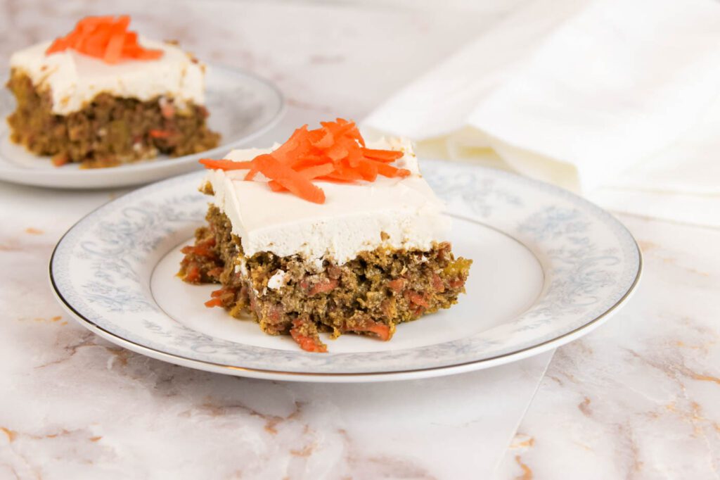 a slice of carrot cake with cream cheese frosting topped with shredded carrots sits on a marble countertop in the kitchen