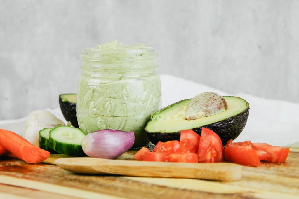half pint jar filled with dressing sitting on a wooden cutting board beside tomatoes, a shallot and a sliced avocado