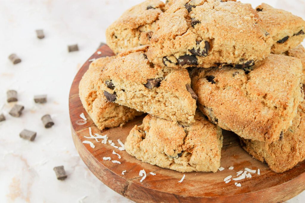 a pile of scones stacked on a wooden stand sits in the forefront with chunks of chocolate in the background