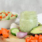 a jar of avocado kefir dressing sits among a bunch of sliced vegetables and a bowl of salad