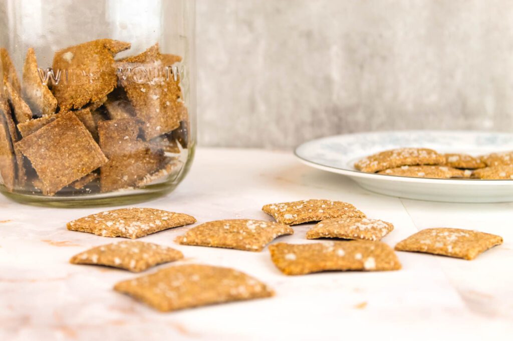 a jar of crackers sits behind a scattering of crackers on a marble countertop