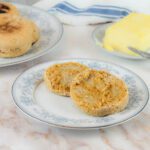 a sliced english muffin with fresh milled flour sits on a plate in front of a plate of butter and a plate full of homemade english muffins
