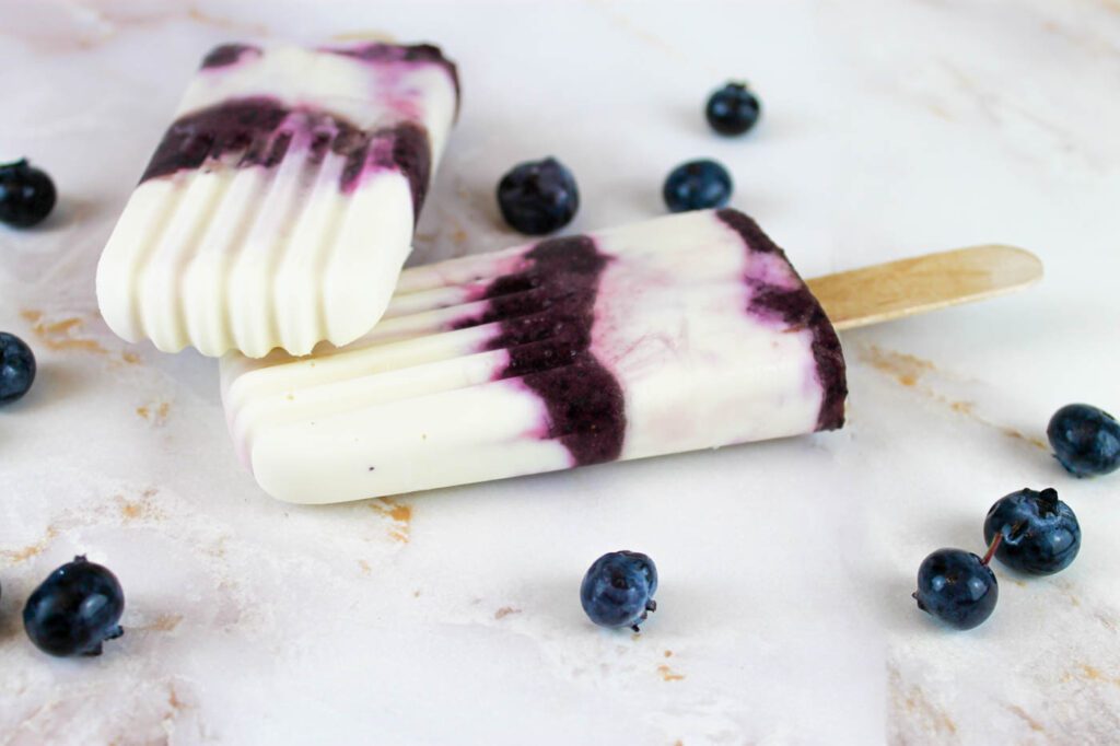 two blueberry cheesecake kefir popsicles lay on top of each other beside scattered blueberries
