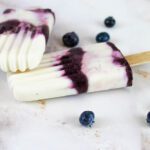 two blueberry cheesecake kefir popsicles lay on top of each other beside scattered blueberries