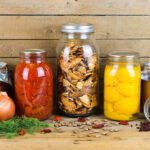 5 glass jars filled with preserved foods sit on a wooden background beside root vegetables and dried food