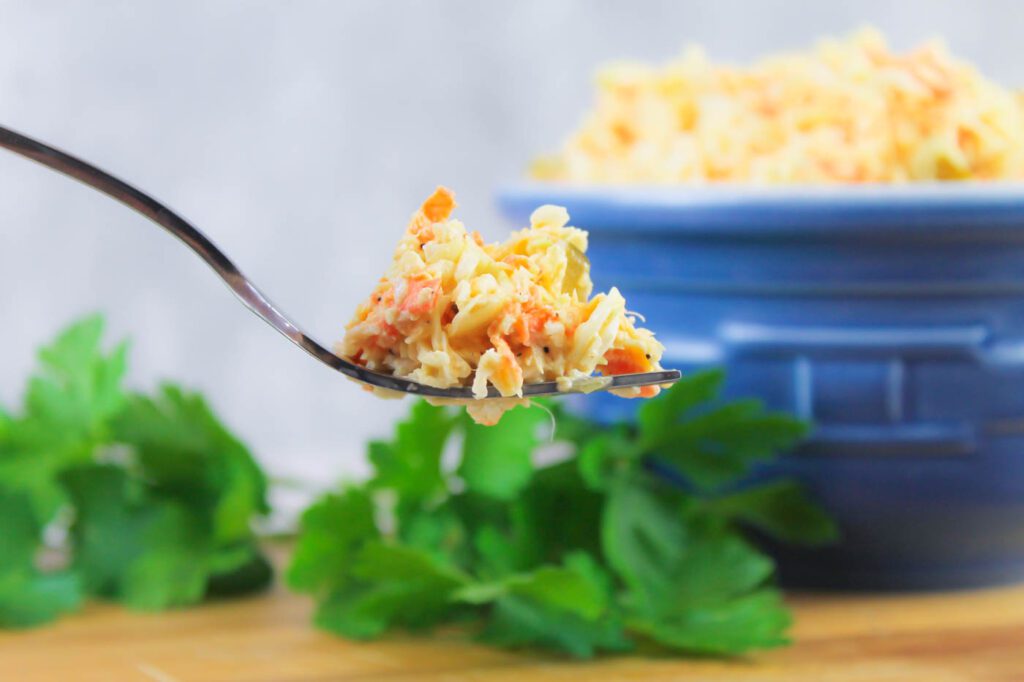 a fork of coleslaw in the foreground with a blue bowl and some fresh herbs in the background