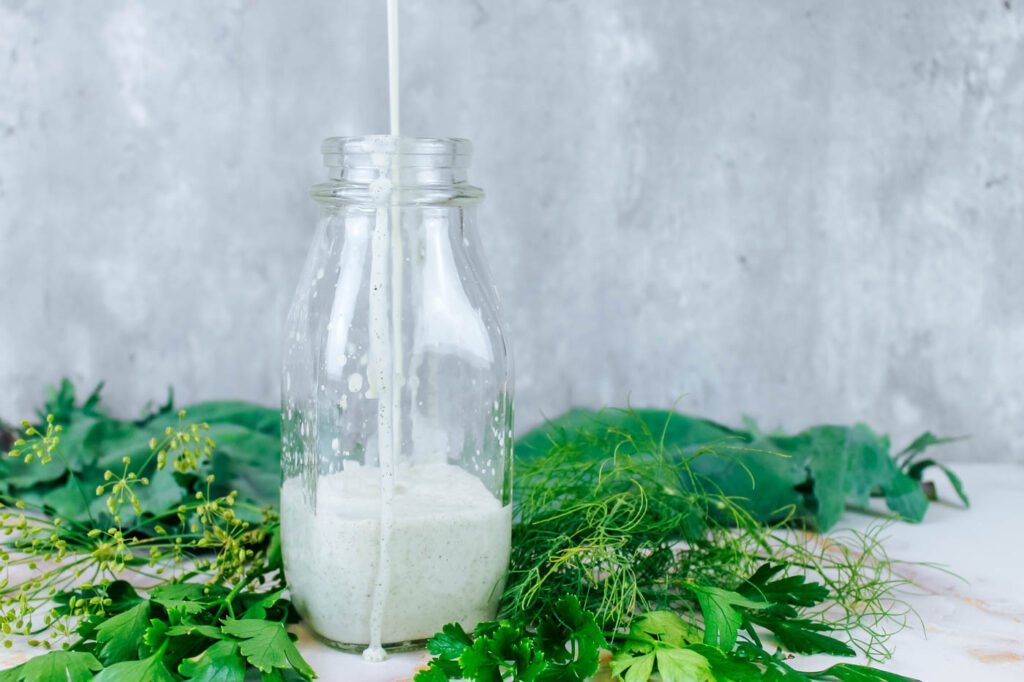 a man pours homemade ranch dressing into a bottle sitting on a countertop next to fresh cut greens